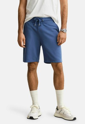 Man draagt blauwe short met trekkoord, witte sneakers, witte sokken, een wit t-shirt en een zilveren polshorloge, staand tegen een effen achtergrond.