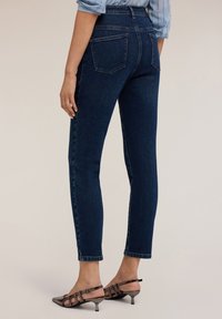 Jeansskiny de mezclilla azul oscuro con cintura alta, con dos bolsillos traseros y detalles de costura a lo largo de las costuras. Llevados con zapatos de tacón bajo.