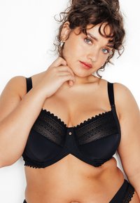 Reggiseno nero con sovrapposizione in pizzo, bordi smerlati e dettaglio decorativo centrale. Tessuto morbido con spalline regolabili.