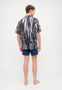 Short-sleeve shirt featuring a black and white abstract pattern, made of lightweight fabric. Paired with navy shorts, model stands barefoot.