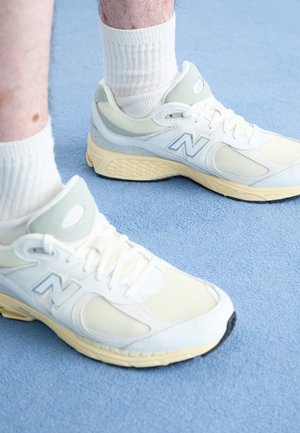 Person iført hvide og beige New Balance-sneakers med hvide ribbede crew-sokker på et blåt tæppebelagt gulv.