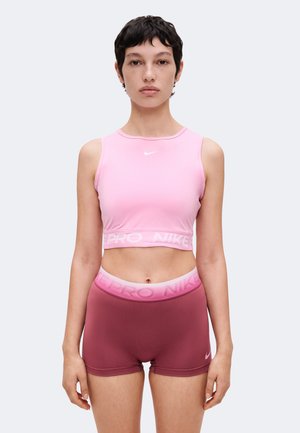 Femme aux cheveux courts et foncés portant un crop top rose Nike et un short Nike Pro bordeaux, debout devant un fond uni clair.
