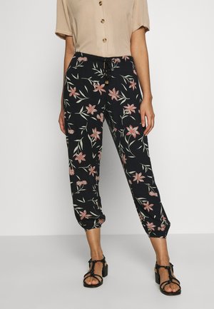 Woman wearing black floral print tapered pants with elastic cuffs, beige short-sleeve buttoned top, and black strappy block heel sandals.