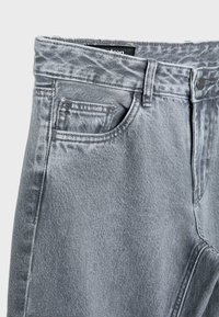Jean en denim gris en gros plan montrant la ceinture, la poche avant avec une petite poche pour la monnaie, la passants de ceinture et la fermeture à bouton métal.
