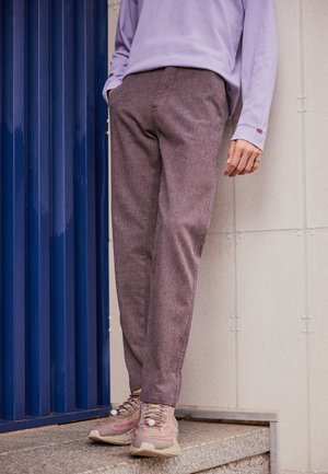 Person iført brune bukser, pink og beige sneakers og en lavendelfarvet langærmet skjorte står nær en blå dør og flisevæg.