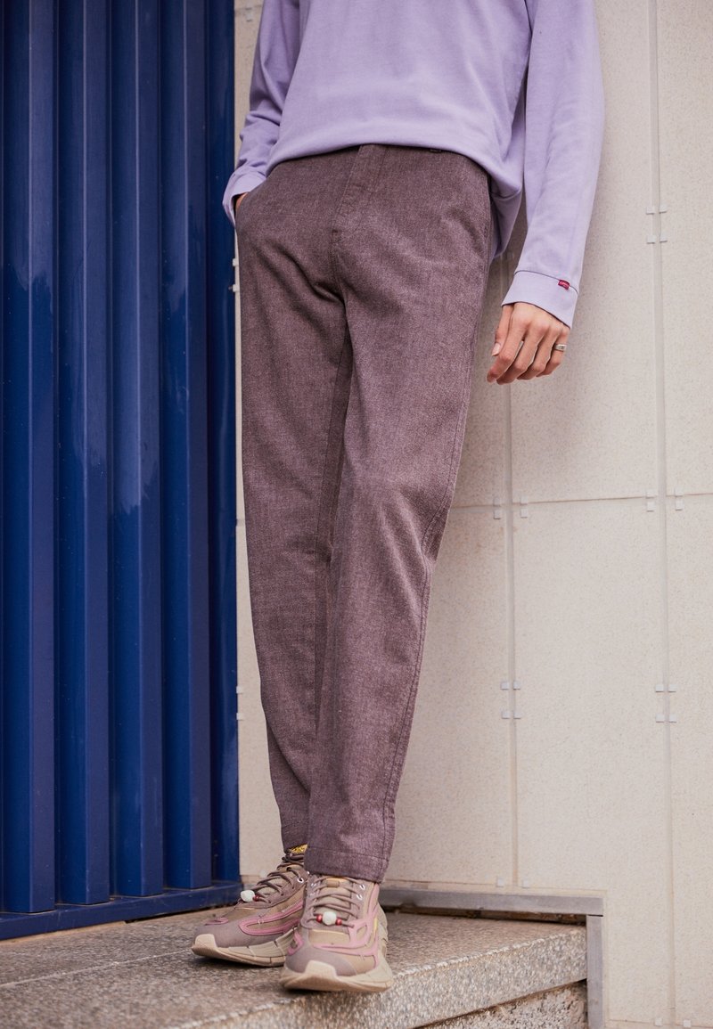 Personne portant un pantalon marron, des baskets roses et beige, et une chemise lavande à manches longues, debout près d'une porte bleue et d'un mur carrelé.