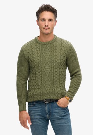 Suéter de punto trenzado color verde oliva con cuello redondo y patrones de diamante texturizados; se lleva con jeans azules y un reloj marrón.