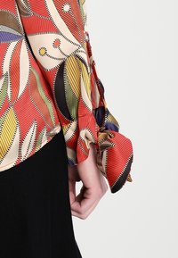 Röd och beige blus med bladmönster, som har detaljer med volangärmar. Slätt tyg med kontrasterande svarta accenter och linjer.