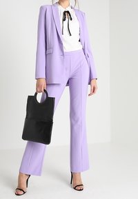 Lila blazer och matchande byxor i kombination med en vit blus med ett svart band, håller en svart tote-väska och bär svarta klackar.