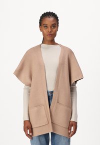 Donna con capelli intrecciati che indossa un gilet di maglia beige aperto davanti su un maglione a coste crema e jeans blu, in piedi di fronte a uno sfondo bianco.