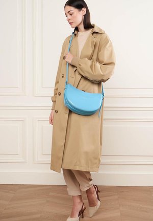 Sac en cuir bleu clair en bandoulière avec une forme arrondie, fermeture éclair et bandoulière adjustable. Porté avec un manteau oversize beige et une tenue neutre.
