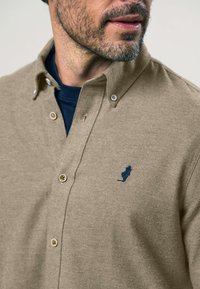 Ein bärtiger Mann trägt ein beiges Hemd mit Knöpfen über einem navyblauen Rundhalsshirt, das ein kleines navyblaues gesticktes Logo auf der Brust hat.