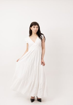 Joven mujer con cabello largo y oscuro, vistiendo un vestido largo blanco sin mangas y tacones negros puntiagudos, de pie contra un fondo claro y sencillo.