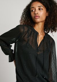 Blusa nera trasparente con tessuto testurizzato e piccole applicazioni, scollatura a V e maniche lunghe, leggermente arricciate.