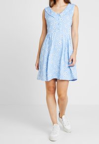 Niebieska sukienka bez rękawów w kwiatowy wzór, z kołnierzykiem, zapięciem na guziki i marszczoną spódnicą. Noszona z białymi trampkami.