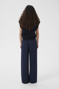 Osoba z długimi kręconymi włosami, ubrana w czarny, bezrękawnik oraz szerokie granatowe spodnie, stoi tyłem na jednolitym białym tle.