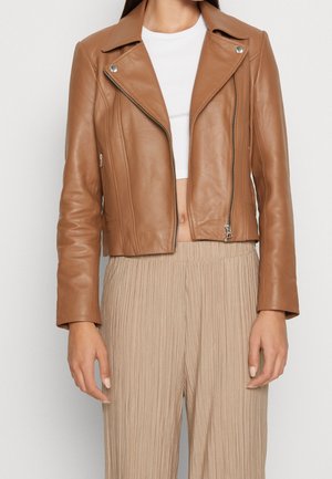 Femme portant une veste de motard en cuir marron clair coupée au-dessus d'un crop top blanc, associée à un pantalon plissé beige taille haute.