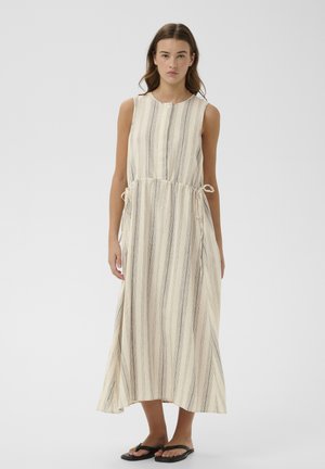 Žena v bezrukávových krémových šatách po členky s vertikálnymi čiernymi pruhmi a bočnými šnúrkami, stojaca pred jednofarebným svetlým pozadím.