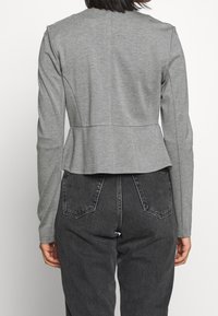 Šedá dlouhá bunda s peplum designem, vyrobená z měkkého materiálu, s kulatým výstřihem a propracovanými detaily na zádech.