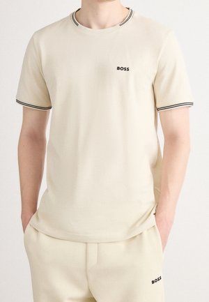 Mann trägt beige kurzärmeliges T-Shirt und passende Hose mit schwarzen "BOSS" Logos und schwarzem Streifenbesatz an Ärmeln und Kragen.