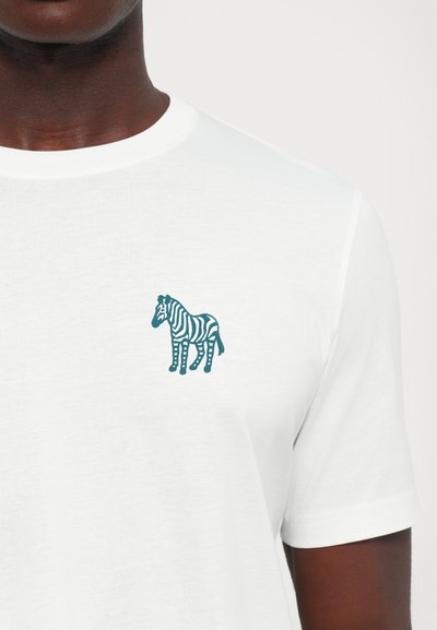 Balta T-kreka ar mazu tumši zilu zebras logo kreisajā krūtī, ko valkā cilvēks ar tumšu ādu.