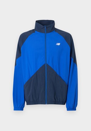 Giacca sportiva blu e blu scuro con zip, colletto alto, polsini elasticizzati e logo New Balance bianco sul petto sinistro.
