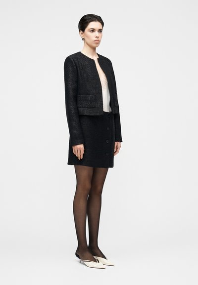 Giacca corta nera con texture, collo tondo e tasche applicate, abbinata a una gonna nera con bottoni e una camicetta neutra. Completano il look collant neri sottili e décolleté bianche.