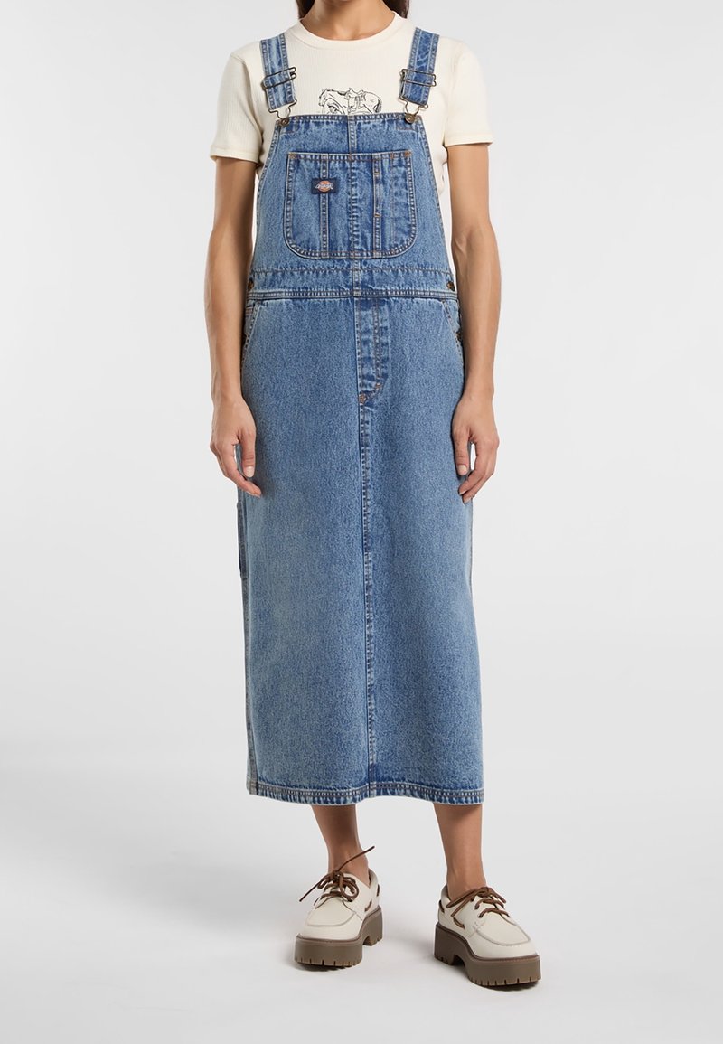 Džínová overall šaty v světle modré, s nastavitelnými ramínky, přední kapsou a volným střihem, spárované se krémovými botami s hnědou podrážkou.