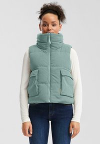 Žena s kučeravými vlasmi, oblečená v svetlom modrom prehrievanom vestu s vysokým golierom, bielej košeli s dlhým rukávom a tmavomodrých džínsoch, stojí čelom.