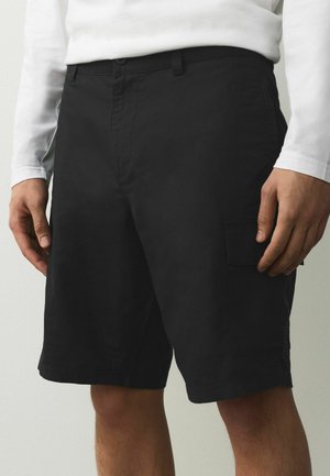Schwarze knielange Cargoshorts mit seitlicher Tasche, getragen mit einem weißen Langarmshirt, zu sehen von der Taille bis zur Mitte des Oberschenkels.