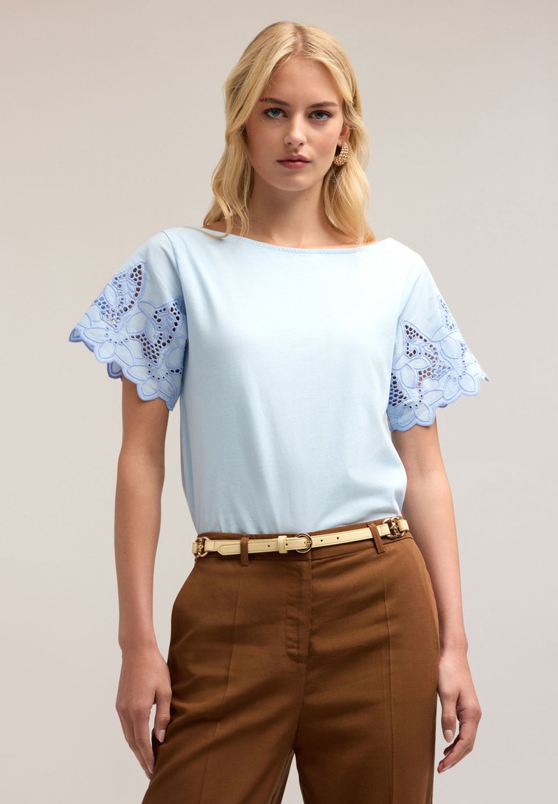Lichtblauw kort mouwtop met geborduurde kantdetails op de mouwen, gecombineerd met een bruine broek en een beige riem.