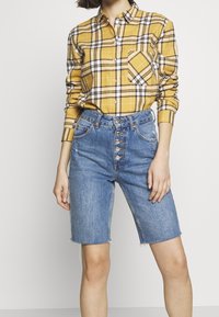 Camicia a quadri gialli con bottoni e maniche lunghe, abbinata a pantaloni corti di jeans blu a vita alta con una chiusura a cinque bottoni e orlo sfrangiato.