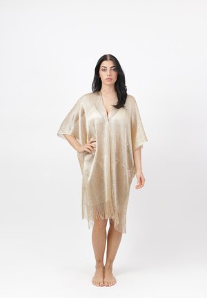 Donna con lunghi capelli scuri in piedi scalza, indossa un abito dorato lucido fino al ginocchio con maniche corte e orlo con frange, mano sul fianco.