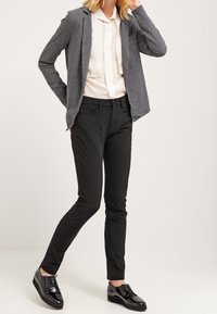 Grå blazer, krämfärgad blus med knytning, svarta slim-fit byxor och glansiga svarta skor. Släta texturer och figursydd silhuett.