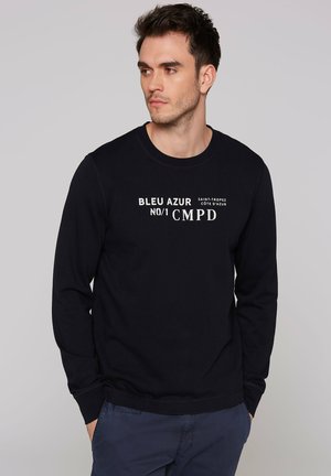 Junger Mann, der nach links schaut, trägt einen schwarzen Pullover mit weißem Text "BLEU AZUR No/1 CMPD" und dunkelblaue Hose vor einem schlichten hellgrauen Hintergrund.