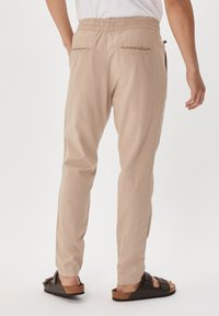 Beige lightweight trousers with an elastic waistband, side pockets, and two back pockets, worn with brown sandals on a plain background.