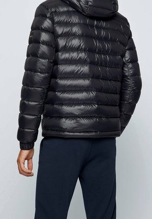 Homme portant une doudoune noire brillante avec capuche, debout dos à la caméra et pantalon bleu foncé sur un fond uni.