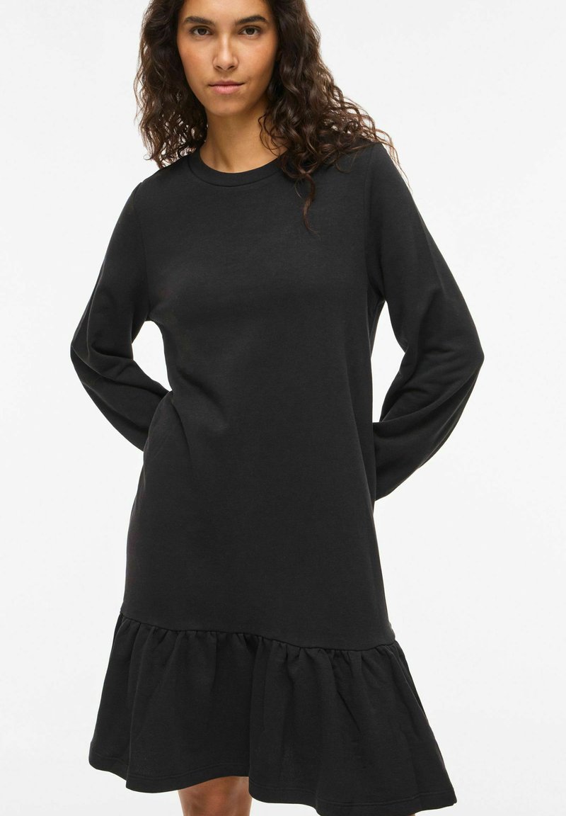 Robe noire à manches longues avec un col rond et un ourlet volanté évasé. Fabriquée en tissu lisse avec une coupe décontractée. Design simple.