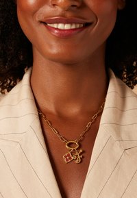 Gouden ketting met een bloemenhanger met rode accenten en een hanger met de letter "F". Het model draagt een lichtgekleurde, gestreepte blazer.