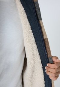 Fliisvooderiga jakk, millel on lukustus, tumedast sinisest välispinnast ja heleda beeži struktureeritud sisepinnaga; nähtavad ruudulised detailid.