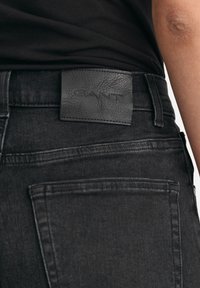 Svarta denimjeans med en läder GANT-etikett på midjan. Har bakfickor med en slät, strukturerad yta.