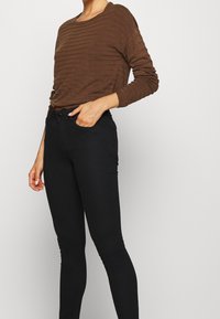 Persona che indossa un top a maniche lunghe marrone e jeans skinny neri a vita alta, con una mano nella tasca anteriore.