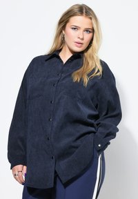 Camicia in corda blu navy con bottoni davanti, due tasche sul petto e maniche lunghe. Presenta bottoni in tonalità argento e una vestibilità leggermente rilassata.