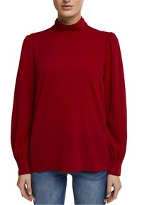 Rød langærmet top med høj halsudskæring, voluminøse ærmer og en glat tekstur; parret med mørke denimjeans.