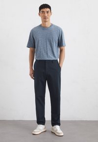 Gestreiftes blaues T-Shirt mit kurzen Ärmeln, kombiniert mit navyblauen Hosen und weißen Turnschuhen mit beigen Akzenten. Einfarbiger Hintergrund.