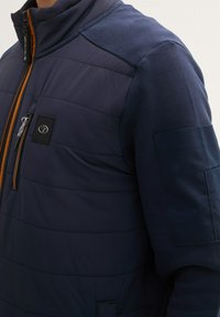 Jachetă de navigație matlasată, cu un buzunar cu fermoar, accente portocalii și panouri contrastante pe umeri. Prezintă un material texturat și detalii netede.