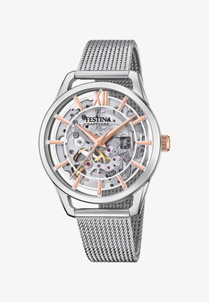 Skeleton hodinky se stříbrným nerezovým pouzdrem, síťovým páskem a průhledným ciferníkem, který odhaluje složité mechanické součásti. Římská číslice XII v růžovém zlatě.