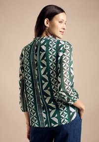 Blusa verde con motivo geometrico bianco. Collo arricciato e maniche a tre quarti. Tessuto morbido e leggero con finitura lucida.