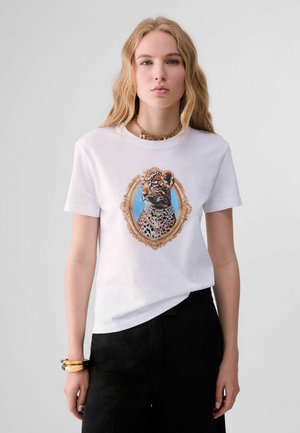 Junge Frau mit langen blonden Haaren trägt ein weißes T-Shirt mit einem Leopardenporträt, das in Gold mit Schmuck gefasst ist, kombiniert mit schwarzen Hosen und goldenen Armbändern.