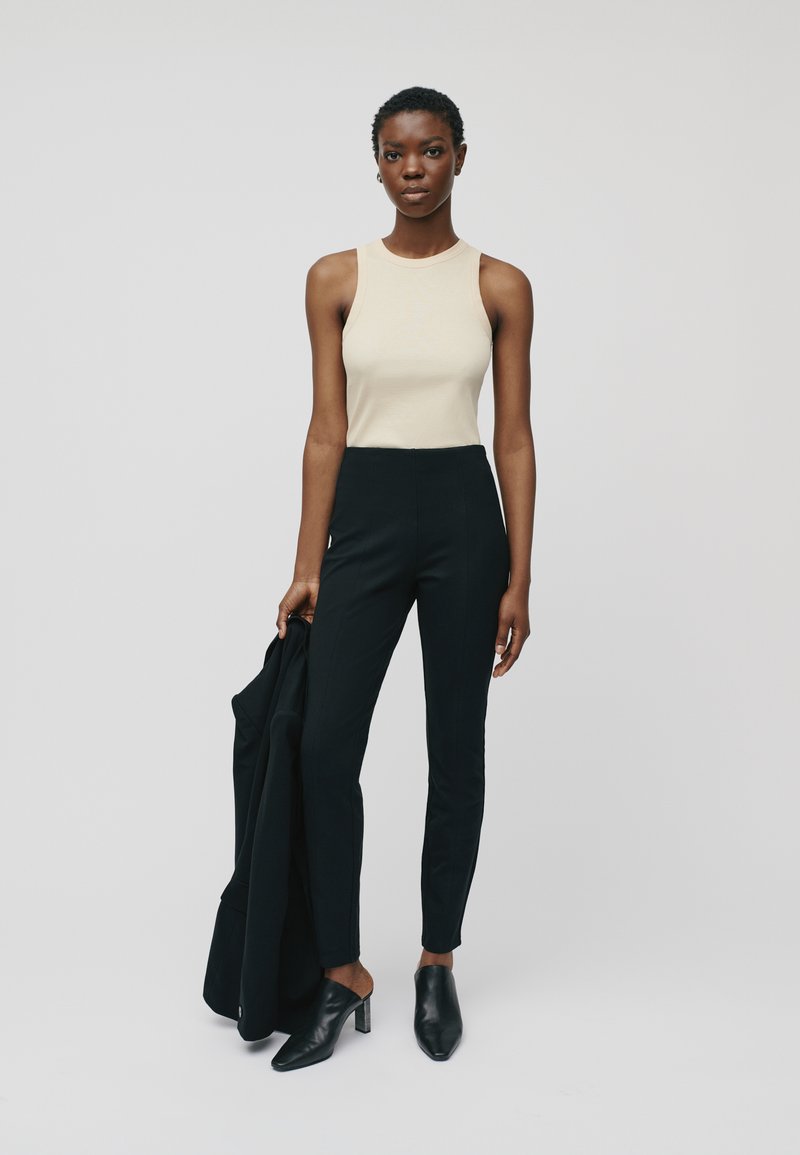 Beige sleeveless top paired with high-waisted black trousers; model holds a black jacket and wears black open-toe heeled mules.
